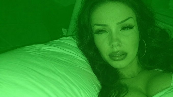A close-up selfie of a woman with long dark hair, dramatic eyelashes, and large hoop earrings, with a green color overlay covering the entire image.