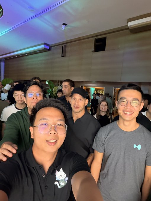 Group selfie of several people posing together at what appears to be an indoor social or networking event, with one person in the foreground wearing a black polo with a Maneki-neko (lucky cat) design.