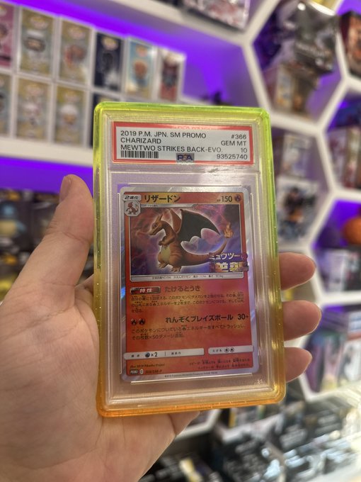 A hand holds a PSA-graded Japanese Charizard Pokémon card labeled '2019 P.M. JPN. SM PROMO CHARIZARD GEM MT 10' from 'MEWTWO STRIKES BACK-EVO.' in front of a display of other cards.