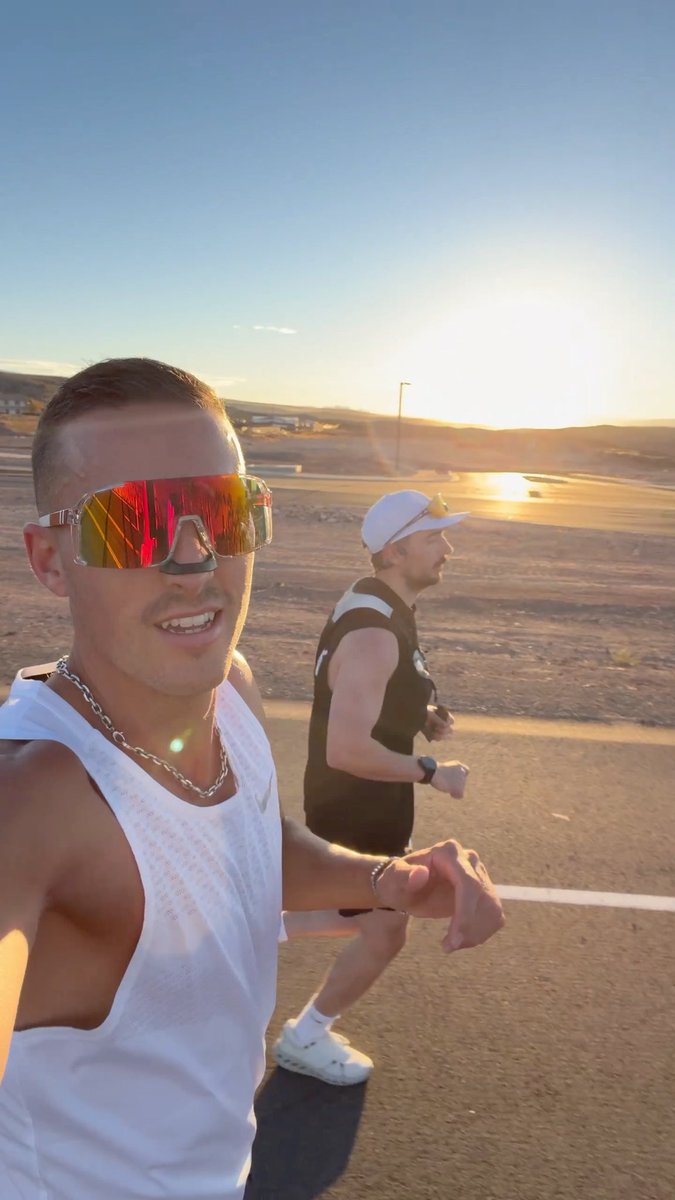 Two men are running on a road at sunset in a desert landscape; the foreground runner wears white sunglasses and a white tank top, taking a selfie video.