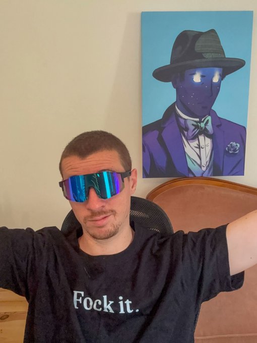 A man wearing sunglasses with reflective blue lenses and a black t-shirt that reads 'Fock it.' sits in front of a painting of a blue figure in a hat.