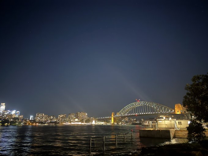 A nighttime photograph of the Sydney Harbour Bridge illuminated against a starry sky, with city lights visible across the harbor.