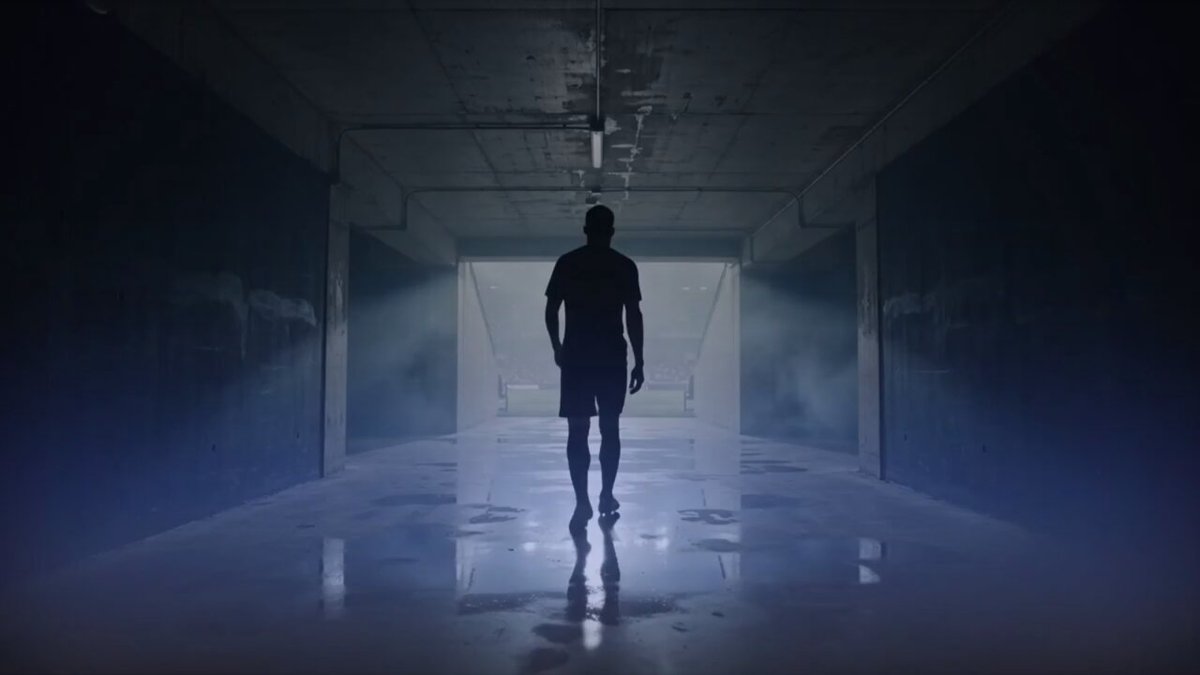 The image shows a man walking through an empty hallway, which appears to be a basement or garage area. The man is wearing shorts and has his back turned towards the camera. There are no other people visible in the photo, making him the main subject of the scene. The background features a series of doors that lead into darkness, creating an atmosphere of mystery and intrigue.