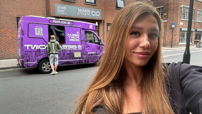 A selfie of a woman with long brown hair standing on a street with a purple Quirk branded van/truck in the background and a 'Black Technology' sign visible.