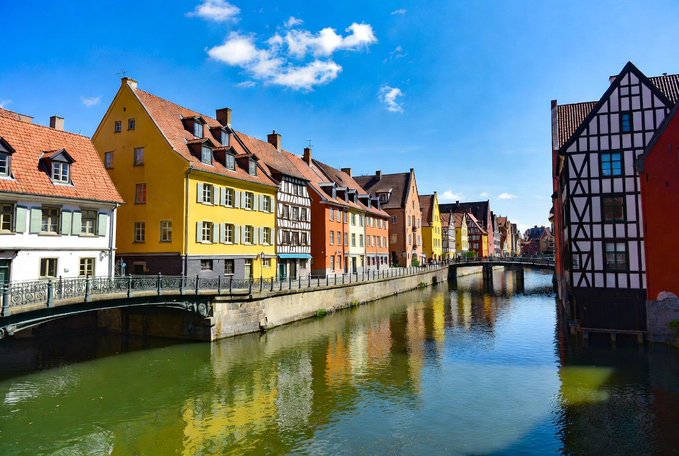 A picturesque canal scene showcases colorful European-style buildings with half-timbered facades in yellow, orange, and white reflecting in the calm water under a blue sky.