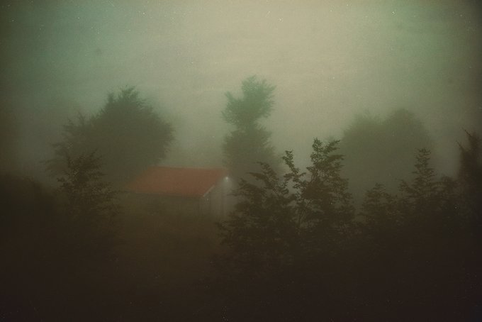 Another dark, foggy photograph showing the same small house with a red roof surrounded by silhouetted trees in heavy mist, nearly identical to the previous image.