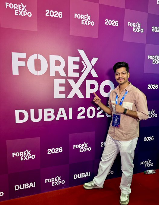 A man poses in front of a pink 'FOREX EXPO DUBAI 2026' backdrop, pointing at the signage while wearing a lanyard with a blue badge.