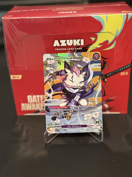 An 'AZUKI TRADING CARD GAME' booster box with a holographic 'Piko of Thousand Blades' card displayed on a stand in front of it.
