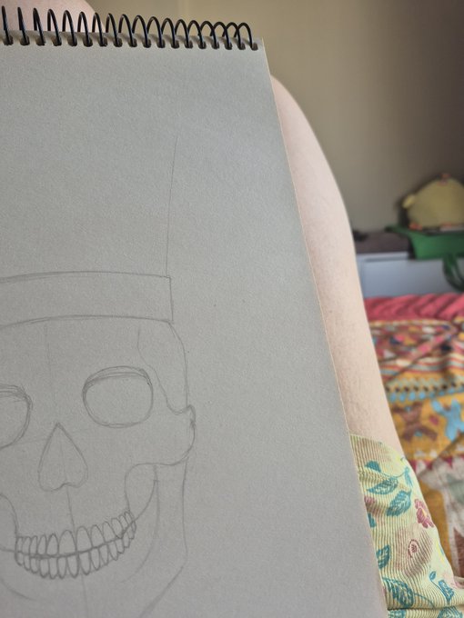 A pencil sketch of a skull wearing a tall hat is in progress on a spiral-bound sketchbook, resting against a floral-patterned surface, capturing the artist's morning creative session with the NFT community.