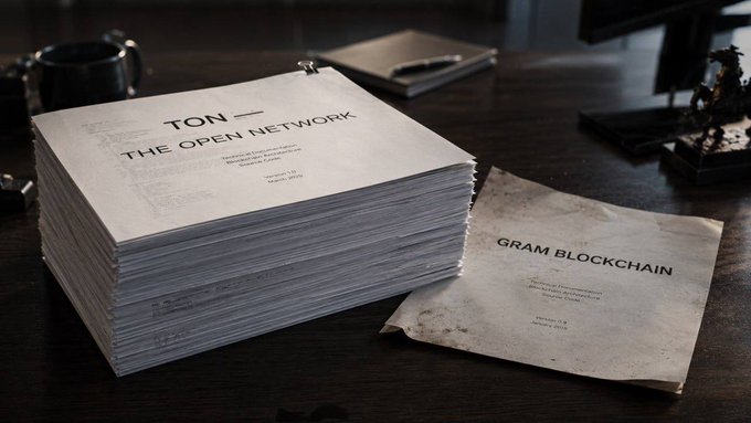 The image shows two stacks of white paper, both with black text that reads "ton network" or "@ton_blockchain", sitting on a desk. The first stack has a book titled "The Open Network" written on top of it, while the second stack has a book titled "Gram Blockchain" written on its cover. Both stacks are placed next to each other and appear to be part of a larger collection or project related to blockchain technology.