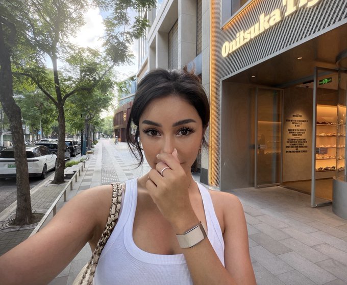 A woman takes a selfie outside an Onitsuka Tiger store, holding her nose as if unable to breathe, cleverly visualizing the post's sentiment about waiting for $ANSEM to return to all-time highs.