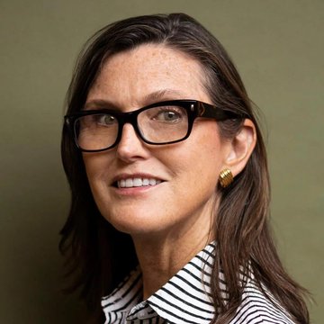 Cathie Wood, CEO of ARK Invest, smiles for a professional portrait wearing glasses and a striped shirt. The image accompanies news of her firm's recent trading activity, including significant purchases of $SPCX and smaller trades in $SOLQ and $HOOD.