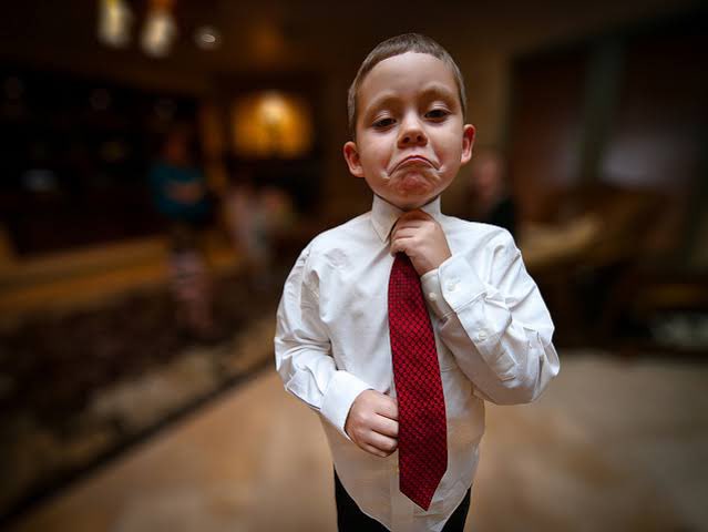 A young boy wearing a white dress shirt and red tie makes a pouting expression while adjusting his collar.