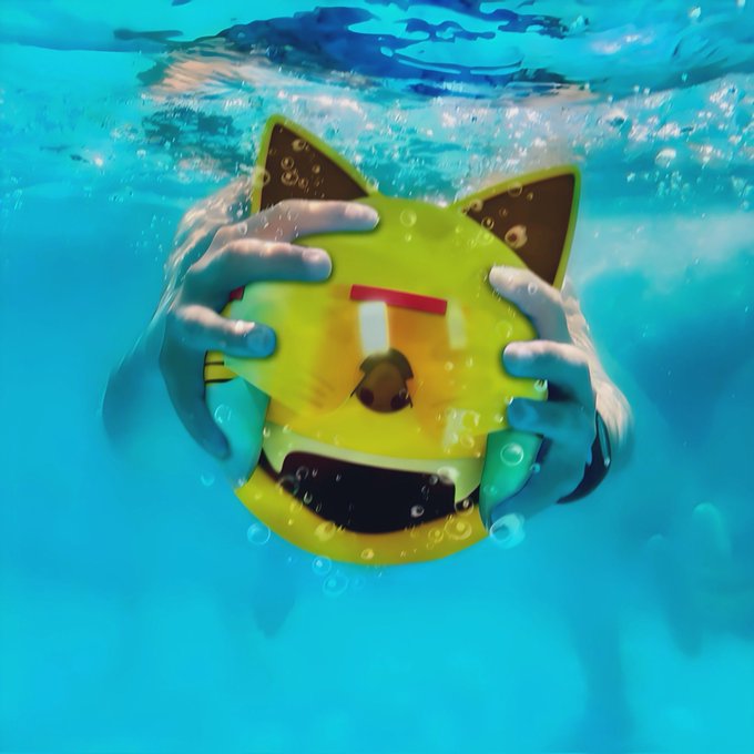 An underwater photograph of hands squeezing a yellow cat-faced plush toy, causing bubbles to rise in the blue water.