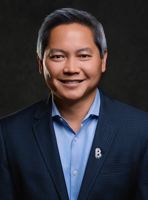 Phong Le, CEO of Strategy, smiles in a professional headshot, wearing a dark checked blazer over a light blue shirt with a "B" pin—likely a Bitcoin symbol—reflecting his role leading the company through strategic BTC trades.
