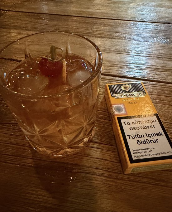 A glass with an ice cocktail and a cherry sits next to a pack of 'COHEX' cigarettes on a wooden table; the packaging features the Greek phrase 'Το κάπνισμα σκοτώνει / Tütün içmek öldürür'.
