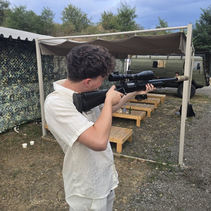 A young man in a cream shirt aims a scoped rifle at an outdoor shooting range.