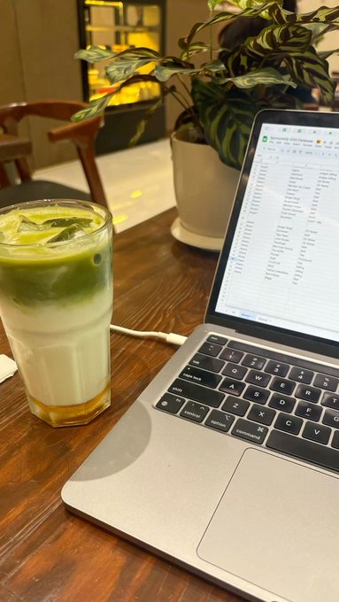 A wooden table scene featuring a glass of iced matcha latte, a MacBook laptop displaying a database interface, and a potted plant in the background of what appears to be a cafe.