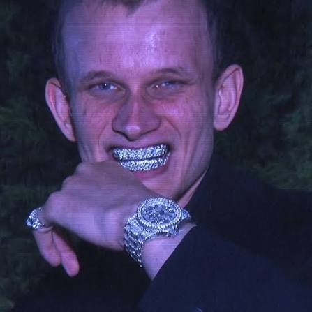 A close-up portrait of a man with a shaved head smiling, showing diamond-encrusted grills on his teeth, wearing a large diamond watch and rings.