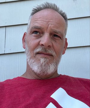 A close-up selfie of a gray-haired, bearded man wearing a red t-shirt, standing in front of white horizontal siding.