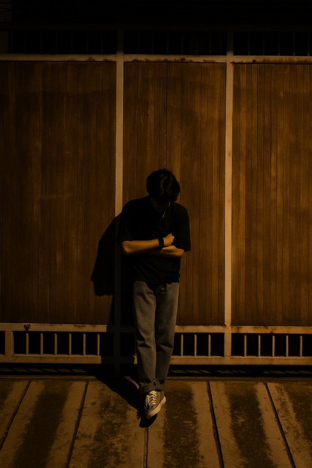 A person in a black t-shirt and jeans leans against a wooden wall at night, head bowed and arms crossed, illuminated by a warm overhead light.