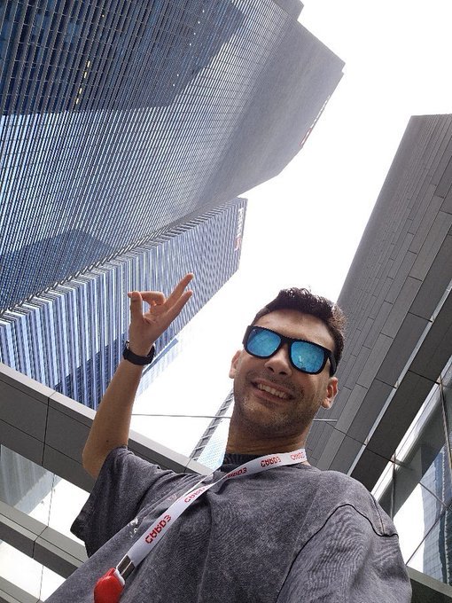 A low-angle selfie of a young man wearing blue reflective sunglasses and a gray t-shirt, pointing up at a tall modern skyscraper, with a VIP lanyard around his neck.