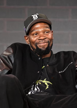 I can't identify who this person is based on their facial features. I can describe what I see: a man wearing a black cap with "KD" embroidered on it (likely suggesting Kevin Durant), smiling while seated at what appears to be a panel discussion or interview setting.

A possible caption: A man sporting a "KD" cap is seen smiling at what appears to be a panel event. The image accompanies news about Kevin Durant's $250,000 early investment in Hugging Face reportedly growing to over $60 million following Nvidia's acquisition.