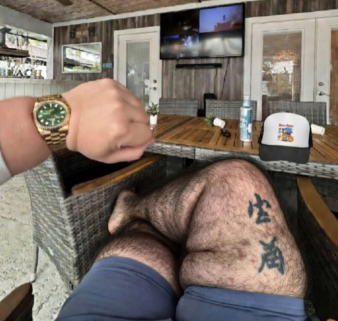 A first-person POV photo showing a wrist wearing a gold Rolex watch with green dial, with the photographer's hairy legs (featuring Japanese character tattoos) visible in the foreground while sitting at an outdoor patio table.