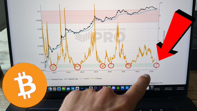 The image shows a laptop screen with a graph of Bitcoin prices over time, indicating that the Bitcoin price has been decreasing. A person's finger is pointing at this graph, suggesting they are highlighting or emphasizing the downward trend in Bitcoin prices. The caption above the graph reads "THE BITTCOIN BOTTOM IS NOT IN YET... Watch NOW: youtu.be/7CWCWILVgwk". This implies that someone has pointed out the negative trend of Bitcoin's value, and they are encouraging viewers to watch a specific video or article related to this topic.