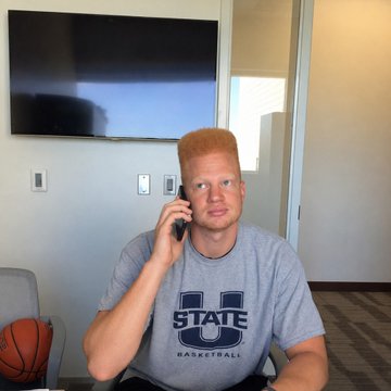 A young man with reddish-blond hair wearing a gray 'U STATE BASKETBALL' t-shirt, talking on a cell phone in a room with a TV mounted on the wall behind him and a basketball on a chair.