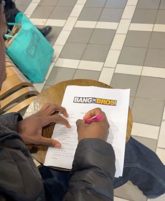 A person fills out a paper titled 'BANG BROS' with a pink pen while sitting at a small wooden table.