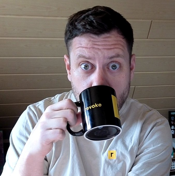 Selfie-style photo of a wide-eyed man drinking from a black 'revoke' branded mug while wearing a matching t-shirt with a small 'r' logo.
