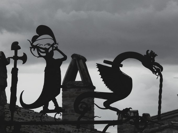 Silhouettes of artistic metal sculptures—a grinning figure with an axe and a dragon-like creature—stand against a dramatic, overcast sky.
