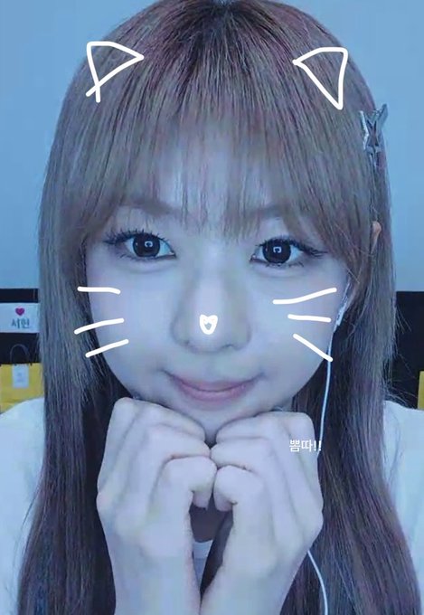 A young woman with long hair and a white top is shown in a close-up photo, with a digital filter applied that adds white cat ears, whiskers, a small heart on her nose, and the text '뿅!!' (Ppyong!!).