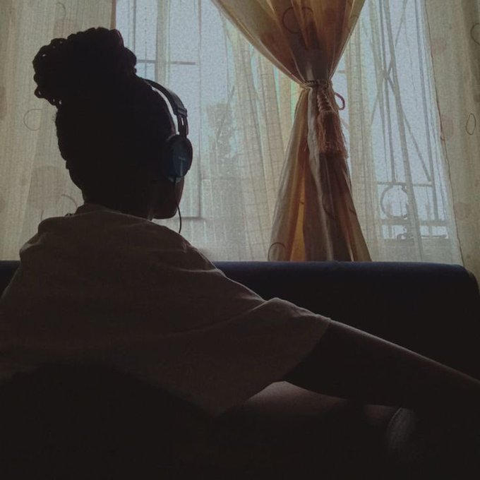 A silhouetted person wearing headphones sits in front of a window with sheer curtains, looking outward.