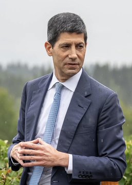 A man with short grey hair wearing a dark blue suit, white shirt, and light blue tie, standing outdoors with a background of green trees.