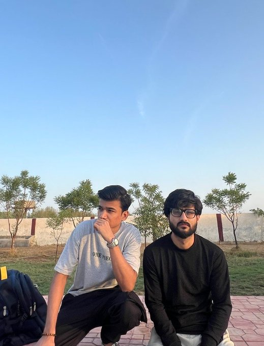 Two young men sit outdoors under a clear blue sky; the one on the left wears a gray "OVENEGY" t-shirt while the other wears a black long-sleeved shirt and glasses.
