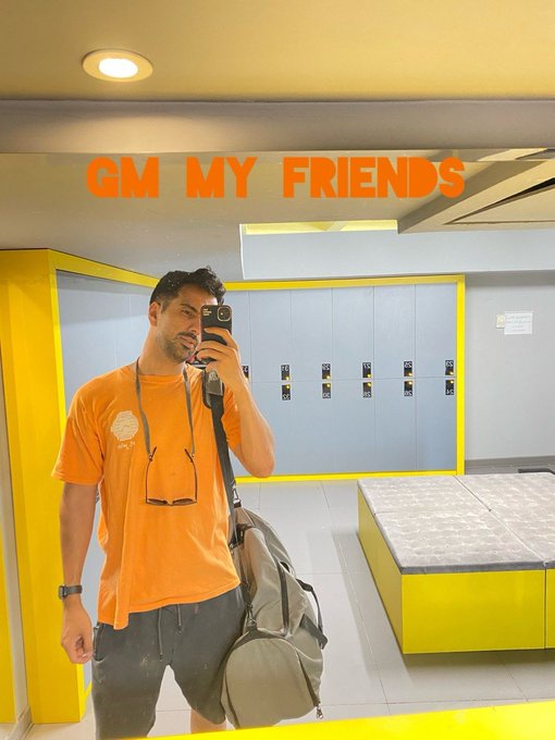 A man in an orange t-shirt takes a mirror selfie with his phone in a yellow-themed room, with the text 'GM MY FRIENDS' overlaid in orange letters.