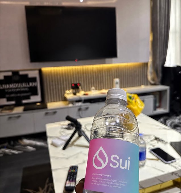 A bottle of Sui blockchain-branded water ('Sui - JOB SUPPLY DRINK - Sui is a next-generation Layer 1 blockchain designed to make digital ownership fast, secure, and accessible for everyone') held in front of a TV in a living room.