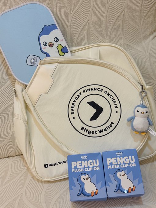 A Bitget Wallet promotional bundle featuring a cream backpack with 'EVERYDAY FINANCE ONCHAIN' logo, a penguin plush clip-on, plush packaging boxes, and a penguin-themed item pouch on a textured background.