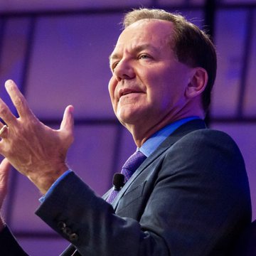 A middle-aged man with light brown hair, wearing a dark suit, blue shirt, and purple tie, speaks animatedly on stage with his hands raised. The image likely depicts billionaire Paul Tudor Jones, referenced in the breaking news about his firm's significant investment in BlackRock's spot Bitcoin ETF.