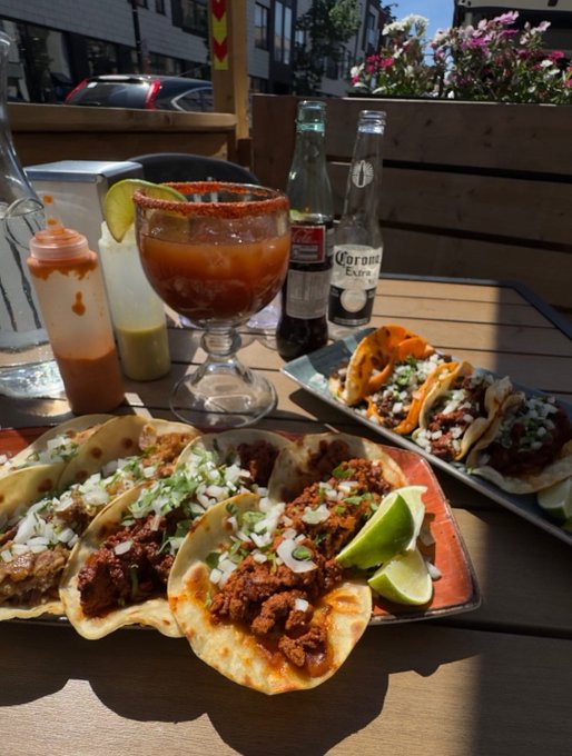 The image features a wooden dining table with several plates of food arranged on it. There are two main dishes prominently displayed - tacos filled with meat and vegetables, as well as burritos stuffed with various ingredients. In addition to these dishes, there is also a plate of nachos topped with cheese and other toppings. 

A bottle of beer can be seen next to the plates, along with a cup on the table. The scene takes place outdoors, possibly at an outdoor restaurant or patio setting. A potted plant adds some greenery to the area, while a car is parked in the background.