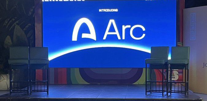 A stage setup featuring a large blue screen displaying 'INTRODUCING Arc' with the Arc logo, flanked by empty teal bar stools on a conference stage.