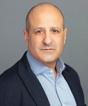 A headshot of a bald man with a light beard, wearing a dark blazer over a light blue button-down shirt, positioned against a gray background. The image accompanies an article about Sharplink CEO Joseph Chalom's comments on Ethereum's role as a global settlement layer compared to Bitcoin.