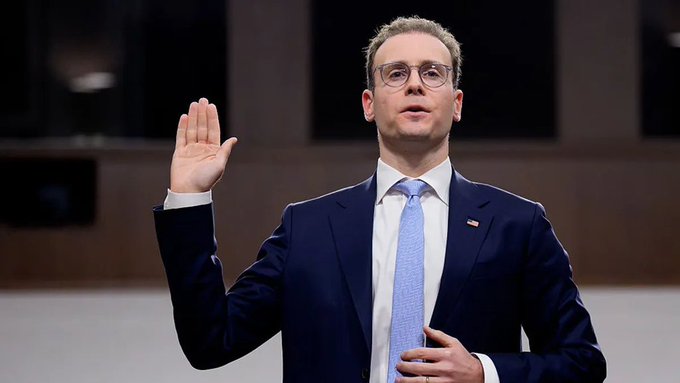 A man in a dark suit, blue tie, and glasses stands with his right hand raised as if taking an oath, wearing a small American flag pin on his lapel.