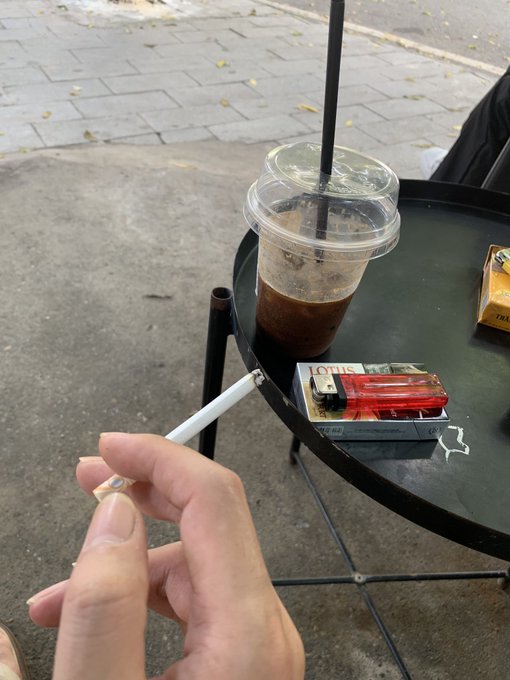 A first-person view of someone holding a cigarette, with a black metal table holding a plastic cup of iced coffee, a red Lotus cigarette pack, and an ashtray by a street curb.