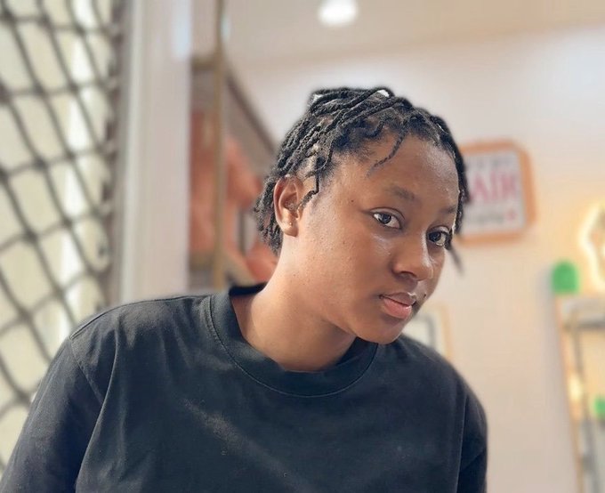A portrait of a young person with short dreadlocks and a black t-shirt, photographed indoors with a softly blurred background.
