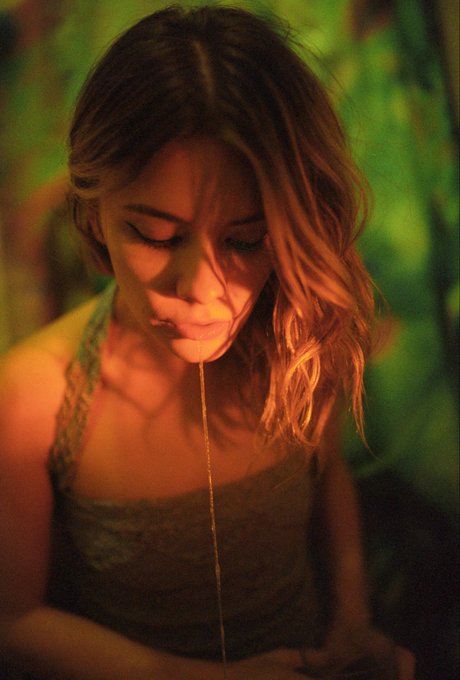 A cinematic close-up of a woman with wavy hair looking downward as a thin stream of liquid pours over her face, illuminated by warm, moody lighting.