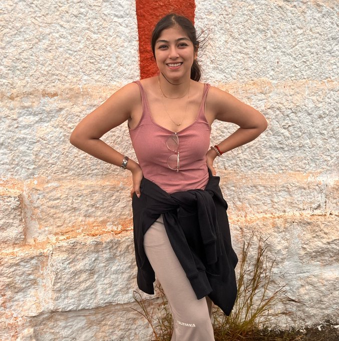 A young woman stands smiling with hands on her hips in front of a stone wall with a red stripe, wearing a pink tank top, beige sweatpants with "ELLESSE" text, and a black jacket tied around her waist. The cheerful, optimistic pose and bright morning greeting pair well with the post's upbeat tone celebrating Bitcoin's return to positive momentum.