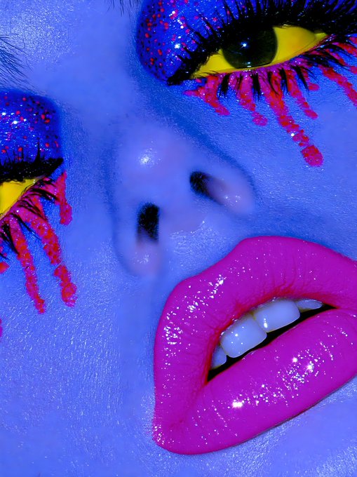 An extreme close-up of a face covered in blue makeup, featuring vibrant magenta lips and yellow eyes accented with pink glittery makeup dripping down the cheeks.